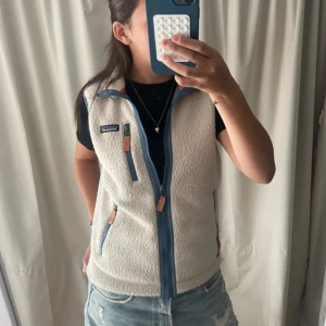 Vit fleeceväst från Patagonia XS - Säljer en vit fleeceväst från Patagonia i storlek XS. Västen har blå dragkedjor, detaljer i orange och tre fickor med dragkedja. Perfekt att slänga över en hoodie eller t-shirt för en chill look. Klassisk Patagonia-logga på bröstet.