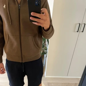 Brun hoodie från Polo Ralph Lauren - Säljer en brun hoodie från Polo Ralph Lauren i storlek M. Tröjan har huva med snörning, dragkedja framtill och två stora fickor. Materialet är mjuk bomull och passformen är normal. Perfekt för chill dagar och snygg att matcha med jeans eller shorts.