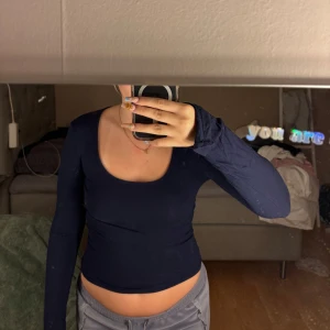 Mörkblå långärmad croppad tröja - Säljer en mörkblå långärmad croppad tröja med snygg djup rundad urringning. Ärmarna har coola tumhål som ger en extra edge till looken. Perfekt att styla med jeans eller cargo pants för en chill vibe. Materialet känns mjukt och stretchigt mot huden.