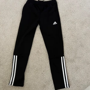 Svarta Adidas träningsbyxor  - Svarta träningsbyxor från Adidas med klassiska vita ränder längs benen och logga på låret. Byxorna har dragkedja vid benslut och resår i midjan med snörning. Tillverkade i polyester, perfekta för sport eller chill. 