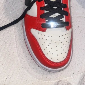 Nike Jordan 1 High röd/svart/vit - Snygga Nike dunk sneakers i rött, svart och vitt skinn. Klassisk siluett med svart swoosh, perforerad tåbox och svarta skosnören. Röd yttersula och coola detaljer som ger en riktigt ikonisk vibe.