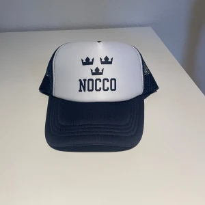 NOCCO X tre kronor keps 🧢  - Snygg truckerkeps från Nocco med vit frontpanel och blått mesh-tyg på sidor och bak. Kepsen har böjd skärm, svart tryck med tre kronor och NOCCO-logga framtill. Perfekt för dig som gillar sportig och clean. Helt ny aldrig använd limiterad 