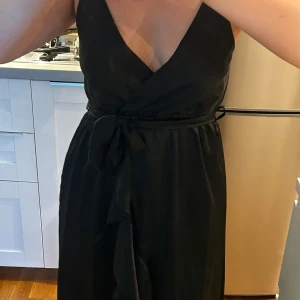 Svart omlottklänning med knytband - Snygg svart omlottklänning med djup v-ringning och knytband i midjan. Klänningen har en omlottdesign med ett knytband i midjan som framhäver figuren. En volangdetalj längs framsidan av kjolen med en slits. Använd någon enstaka gång. Köpt utomlands så det finns ingen storlek men jag är S. Skulle säga att det passar även XS och M.
