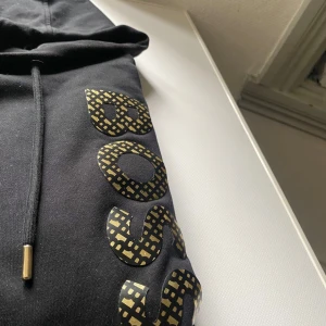 Svart hoodie från BOSS med guldtryck - Svart hoodie från BOSS med stor huva och snörning med guldfärgade detaljer. Snyggt BOSS-tryck i guld med rutigt mönster längs ena sidan. Tillverkad i merceriserad bomull som känns mjuk och lyxig mot huden. Perfekt för dig som gillar stilrena streetwear-plagg.