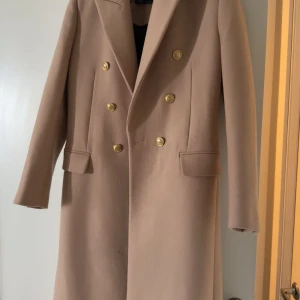 Beige kappa med guldknappar - Snygg beige kappa med dubbelknäppning och guldiga knappar framtill. Klassisk krage och två fickor med lock. Rak och stilren modell som ger en clean look. Perfekt för dig som gillar tidlös stil och vill sticka ut med detaljer. Nypris 1699