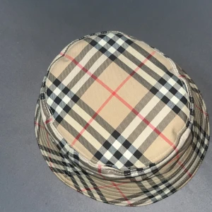 Burberry rutig bucket hat beige - Snygg bucket hat från Burberry i klassiskt beige rutigt mönster med svarta, vita och röda detaljer. Hatten har bred brätte och är tillverkad i bomull, perfekt för att lyfta din stil med en ikonisk touch. Den är strl L använd i cirka en vecka tar byten och pris kan diskuteras 
