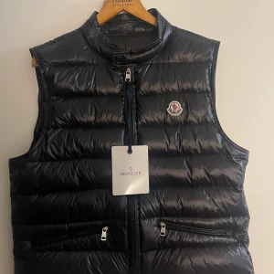 Svart dunväst från Moncler - Snygg svart dunväst från Moncler med glansig finish och klassisk logga på bröstet. Västen har hög krage, två dragkedjefickor och stängs med dragkedja framtill. Perfekt för lager-på-lager och streetstyle.