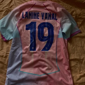 Barcelona Lamine Yamal tröja - Säljer en FC Barcelona fotbollströja med Lamine Yamal och nummer 19 på ryggen. Tröjan har coola färgblock i rosa, ljusblått och lila, med tunna vertikala ränder och detaljer i turkos. Nike-logga, Spotify-sponsor och FCB-emblem på bröstet. Materialet är lätt polyester.