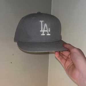 Grå LA-keps från New Era - Snygg grå keps från New Era med LA-logga broderad framtill i vitt. Klassisk modell med rak skärm och broderade ventilationshål. MLB-logga baktill och New Era-logga på sidan. Perfekt för dig som gillar streetwear och sportig stil. Storleken är 7 1/4 eller 57,7cm
