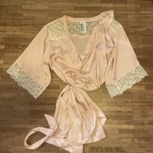Ljusrosa morgon rock  - Superfin ljusrosa rock från H&M med breda spetsdetaljer vid axlar och ärmslut. Klänningen har knyt i midjan och är tillverkad i mjukt, glansigt material som ger en lyxig känsla. Perfekt för dig som gillar romantisk och feminin stil.