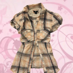 Rutig kortärmad skjorta i beige och blå - Kortärmad skjorta från F&F med rutigt mönster i beige, blått och orange. Skjortan har puffiga ärmar, två bröstfickor och knappar framtill. Perfekt för dig som gillar en avslappnad men ändå snygg stil.