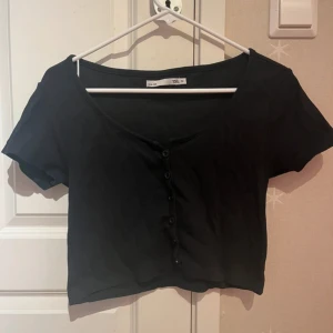 Svart croppad topp med knappar - Svart croppad topp med korta ärmar och v-ringning. Toppen har knappar framtill och är i mjukt material, perfekt att styla med jeans eller kjol. Enkel och stilren design som funkar till många olika looks.