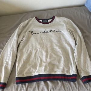Grå sweatshirt från Bondelid, M - Snygg grå sweatshirt från Bondelid med broderad logga på bröstet. Tröjan har ribbade muddar och nederkant med mörkblå och röda ränder. Rund halsringning med samma randiga detalj. Perfekt för chill dagar och enkel att matcha med jeans.