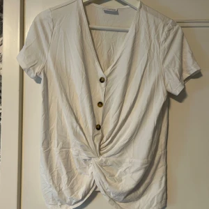 Vit blus med dekorativa knappar - Vit kortärmad topp med v-ringning och snygg omlott/knytdetalj framtill. Tre dekorativa knappar i trälook ger en cool touch. Toppen har twistad front, perfekt för en chill och stilren look.