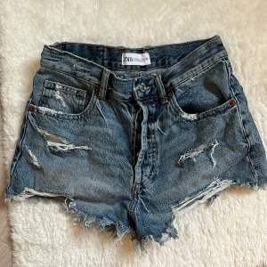 ZARA Blå jeansshorts med slitningar  - Snygga blå jeansshorts från Zara slitna detaljer framtill och baktill. Klassisk femficksmodell med knapp och dragkedja. Perfekt för dig som gillar en edgy och avslappnad stil.