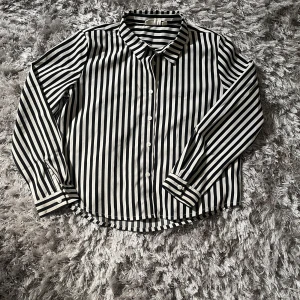 Randig skjorta från KappAhl - Snygg skjorta från KappAhl med svartvita vertikala ränder och klassisk krage. Skjortan har långa ärmar, knappar framtill och cool detalj med diagonala ränder på ryggen. Perfekt för dig som gillar en stilren och trendig look.