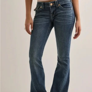 True Religion blå bootcut jeans - Snygga blå jeans från True Religion med bootcut passform och klassiska kontrastsömmar. Jeansen har ikoniska bakfickor med gul brodyr och lock, samt guldiga knappar. Tillverkade i mjukt denim med en lätt tvättad look. Perfekta för dig som gillar en tidlös och cool stil.