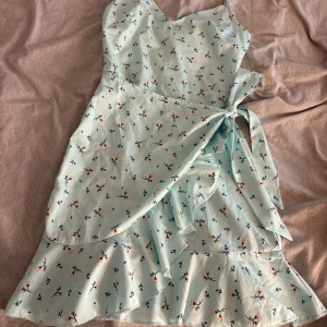 Ljusblå blommig omlottklänning - Superfin ljusblå kort omlottklänning med små blommor i orange och mörkblått. Klänningen har smala axelband, volangkant nertill och knyt i midjan. Perfekt för varma dagar och ger en söt och somrig vibe.