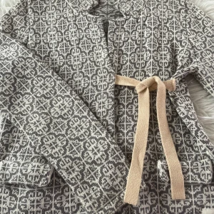 Mönstrad cardigan jacka Odd Molly - Superfin cardigan jacka från Odd Molly i grått och beige med stickat mönster och snyggt knytband i midjan. Jackan har långa ärmar, fickor fram och små rosetter bak. Perfekt för dig som gillar unika detaljer och mysig stil. Den är i storlek 2 och motsvarar ungefär strl S! Har inga tecken på användning!