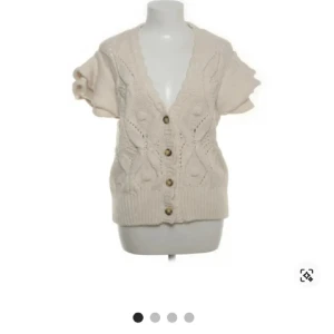 Beige stickad kofta med kort ärm - Superfin beige stickad kofta med korta ärmar och snyggt mönster framtill. Koftan har v-ringning och knappar hela vägen ner, samt ribbad kant nedtill. Perfekt att styla med jeans för en chill look.