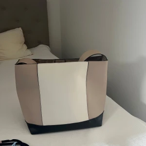 Beige och vit axelväska i skinnimitation - Stilren axelväska i beige, vitt och svart ett rejält handtag. Väskan har ett stort huvudfack, innerficka med dragkedja och magnetknäppning upptill. Väskan är från Lindex och är näst intill oanvänd