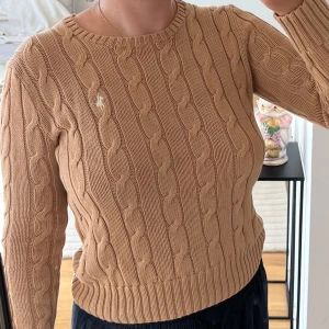 Kabelstickad tröja från Ralph Lauren - Snygg beige kabelstickad tröja från Polo Ralph Lauren i mjuk bomull. Klassisk rund halsringning, ribbade muddar och långärmad modell. 