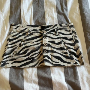 Zebramönstrad kortkjol från Lindex - Snygg kortkjol från Lindex med zebramönster i svart och beige. Kjolen har fyra framknappar, bälteshällor och både fram- och bakfickor. Materialet är jeansliknande bomull som ger en cool och edgy vibe. Perfekt för dig som gillar att sticka ut!