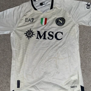 Vit Napoli fotbollströja EA7 Small - Napoli fotbollströja i vitt från EA7, officiell special edition. Tröjan har korta ärmar, svarta detaljer och klubbmärke på bröstet. MSC och Upbit-loggor samt italienska mästarmärket framtill. Tillverkad i lätt och ventilerande material, perfekt för match eller träning.