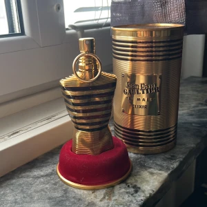 Jean Paul Gaultier Le Male Elixir - Exklusiv parfym i metallisk guldflaska formad som en manlig torso, med svarta ränder och detaljerad design. Kommer i en lyxig guldfärgad burk med märkets logga och röd sammetsbas. Perfekt för dig som gillar statement-dofter och snygg inredningsdetalj.
