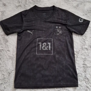Svart Borussia Dortmund fotbollströja Puma - Snygg svart Borussia Dortmund fotbollströja från Puma med diskret mönster och silvriga detaljer. Tröjan har klubbmärke, sponsorlogga och två stjärnor på bröstet samt korta ärmar och V-ringning. Tillverkad i lätt och ventilerande material. Passar S och M