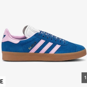 Adidas Gazelle blå/rosa sneakers - Snygga Adidas Gazelle sneakers i blå mocka med ljusrosa detaljer och klassiska tre ränder. Skon har en brun gummisula och snörning framtill. Perfekt för dig som vill sticka ut med färg och stil.