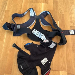 Svart mesh bärsele från BabyBjörn - Säljer en svart bärsele från BabyBjörn i luftig mesh. Selen har justerbara remmar och tydliga instruktioner för användning. Perfekt för att bära små barn nära kroppen och den har en liten säck som är enkel att packa ihop allting i. Snygg och praktisk design med smarta detaljer.