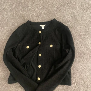 Svart cardigan jacka från H&M med guldknappar - Stilren svart cardigan jacka från H&M med snygga guldknappar framtill. Jackan har rund halsringning, två dekorativa fickor och är tillverkad i ett mjukt, stickat material. Perfekt för dig som gillar klassiska detaljer och vill ha en jacka som sticker ut.