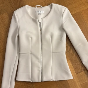 Vit kavajjacka från Amisu, strl 34 - Snygg vit kavajjacka från Amisu i figurnära modell med dragkedja framtill. Jackan har rund halsringning, långa ärmar och markerade sömmar som ger en clean och stilren look. Perfekt för dig som gillar minimalistisk stil.