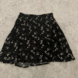 Svart blommig kjol från Xoxo Friend - Säljer en svart kortkjol från Xoxo Friend med gulligt blommigt mönster i vitt och ljusgult. Kjolen har en lätt utställd form och är gjord i mjukt, tunt tyg. Perfekt för dig som gillar en söt och avslappnad stil.