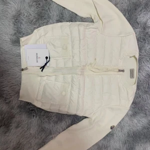  Moncler cardigan  - Snygg vit dunväst från Moncler med quiltad framsida och två stora fickor med knappar. Västen har dragkedja framtill och Moncler-logga på ärmen. Perfekt för lager-på-lager och stilren look.