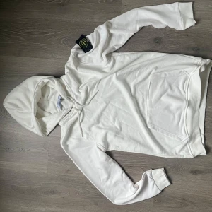 Vit hoodie från Stoneland XXL - Snygg vit hoodie från Stoneland i storlek XXL. Klassisk modell med huva, snörning och stor magficka. Mjuk bomullskänsla och cool svart logotyp-patch på vänster ärm. Perfekt för dig som gillar enkel och clean stil.