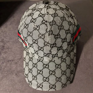 Gucci keps med GG-mönster - Exklusiv keps från Gucci i grått och svart med klassiskt GG-monogram över hela kepsen. Snygga gröna och röda ränder på sidorna. Tillverkad i merinoull och kashmir för en lyxig känsla. Justerbar baktill för perfekt passform.