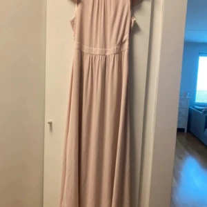 Ljusrosa långklänning med ärmlös design - Säljer en elegant ljusrosa långklänning med ärmlös design och rundad hals. Klänningen har ett markerat midjeband och ett mjukt, flytande fall. Perfekt för dig som gillar stilrena och feminina plagg. Materialet känns lätt och bekvämt mot huden.