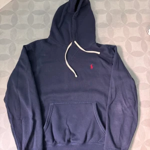 Polo Ralph Lauren Hoodie Storlek S - Snygg mörkblå hoodie från Polo Ralph Lauren med klassisk röd logga broderad på bröstet. Tröjan har huva med vita snören och en stor magficka. Perfekt för dig som gillar en clean och stilren look.