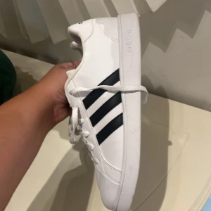 Vita Adidas sneakers med svarta ränder - Klassiska vita Adidas sneakers med svarta ränder på sidorna och vit sula med hexagonmönster undertill. Skorna har snörning och är tillverkade i syntetmaterial med meshdetaljer på plösen. Perfekta för en clean och sportig look. 
