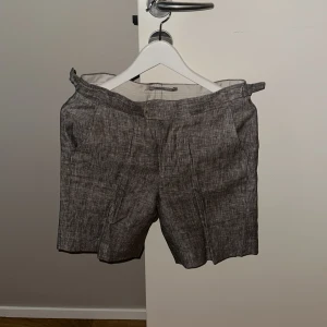 Grå linneshorts från John Henric - Snygga grå dress-shorts från John Henric i linne med diskret melerad look. Modellen är med justerbara spännen i midjan och har både fram- och bakfickor med knappdetaljer. Perfekta för chill dagar under sommaren där man både kan klä upp dem och klä ner dem.