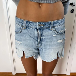 Ljusblå ripped jeansshorts - Snygga ljusblå jeansshorts med slitna detaljer och fransig kant. De har klassisk femficksdesign, låg midja och coola revor både fram och bak. Perfekta för en chill och avslappnad stil under varma dagar.