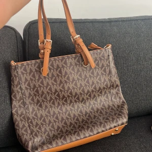Brun axelväska från Michael Kors - Snygg brun axelväska från Michael Kors med klassiskt MK-mönster över hela väskan. Väskan har justerbara remmar i ljusbrunt skinn och detaljer i guld. Perfekt storlek för att få plats med allt du behöver till vardagen.