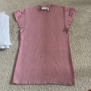 Rosa ribbad topp från Vila - Supersöt rosa topp från Vila med ribbad struktur och korta ärmar. Ärmarna har utskurna blommönster som ger en extra detalj. Perfekt för dig som gillar en feminin och trendig stil.