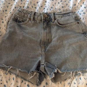 Ljusblå jeansshorts från BikBok - Snygga ljusblå jeansshorts från Never Denim i modell midwaist. Klassisk femficksdesign, råa fransiga kanter och avslappnad passform. Perfekta för sommardagar och lätta att matcha med allt. Styla med en crop top eller hoodie för en chill vibe.