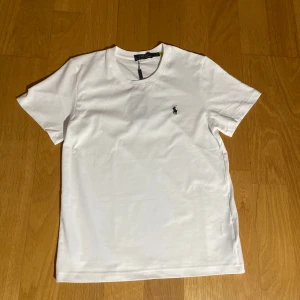 Vit t-shirt från Polo Ralph Lauren - Klassisk vit t-shirt från Polo Ralph Lauren med rund hals och korta ärmar. T-shirten har den ikoniska broderade loggan på bröstet och är gjord i mjuk bomull. Perfekt basic-plagg med snygg passform och stilren look. storlek S passar en som är 164cm