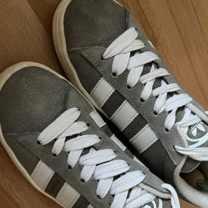 Adidas Campus grå/vita sneakers stl 40 - Snygga Adidas Campus sneakers i grå mocka med klassiska vita ränder och vit logga på hälen. Skorna har vit sula, vita skosnören och en stilren, retro vibe. Perfekta för dig som gillar streetstyle och vill ha något som sticker ut men ändå är enkelt att matcha.