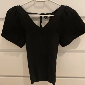Svart ribbad topp med puffärm och knyt - Svart ribbad topp med bred v-ringning från only och puffiga korta ärmar. Snygg knytning i ryggen som ger en extra detalj. Stretchigt material som sitter slim och framhäver figuren. Perfekt för dig som gillar statement-ärmar och stilren look.