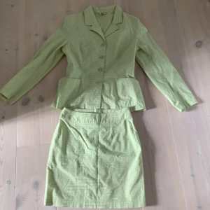 Grön rutig kavaj och kjol från Flash - Snyggt tvådelat set från Flash med kavaj och kjol i ljusgrönt rutigt mönster. Kavajen har klassisk krage, knappar framtill och markerad midja. Kjolen är rak och höftlång. Perfekt för dig som gillar retrovibbar och pastelliga färger.